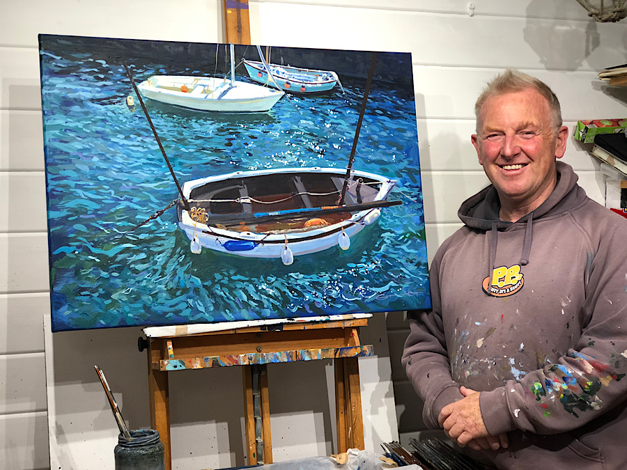 Artist Steve PP stood next to an easel with a painting of boats in a harbour on a sunny day on it